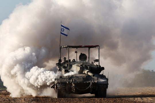 Een Israëlische tank, met Israëlische vlag, rijdt langs de grens met de Gazastrook omgeven door een grote rookpluim.