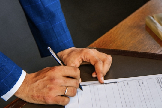 Foto van de handen van iemand in een blauw kostuum die documenten ondertekent.