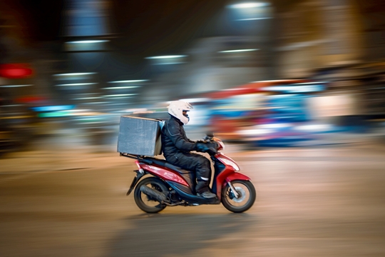 Een maaltijdkoerier rijdt op een scooter door een stedelijke omgeving, de achtergrond is zo geblurd dat het lijkt alsof de scooter stilstaat en de omgeving zeer snel beweegt.